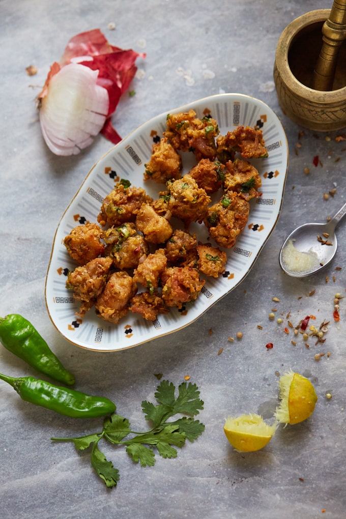 A plate of Chicken pakora surrounded with green chillis, lemon, onion and coriander seeds.