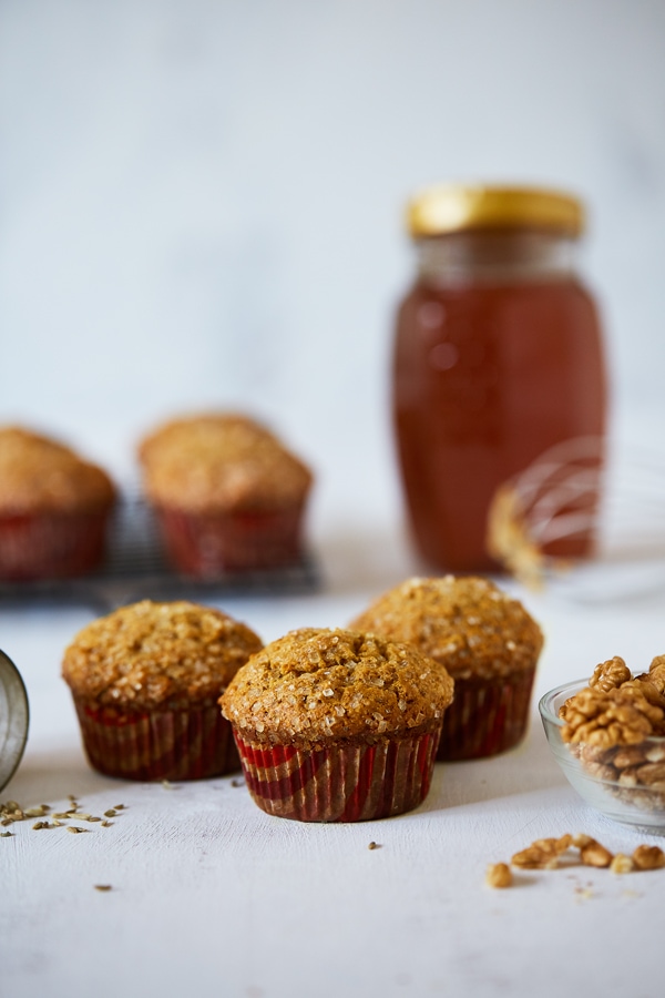 Honey Cornbread Muffins (Makki Muffins) on a table with fennel seeds and walnuts, in the background is a jar of honey, a whisk and more muffins on a cooling rack.