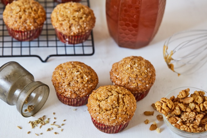 Honey Cornbread Muffins (Makki Muffins) surrounded by fennel seeds, walnuts, a jar of honey, a whisk, and a cooling rack of more muffins.